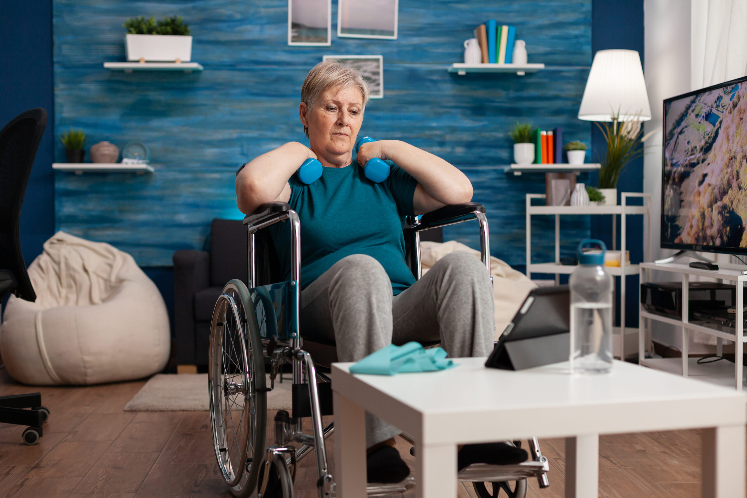 Invalid senior woman in wheelchair watching gym body exercise on tablet in living room exercising arms muscle using workout dumbbells. Pensioner recovery after paralysis training muscles resistance